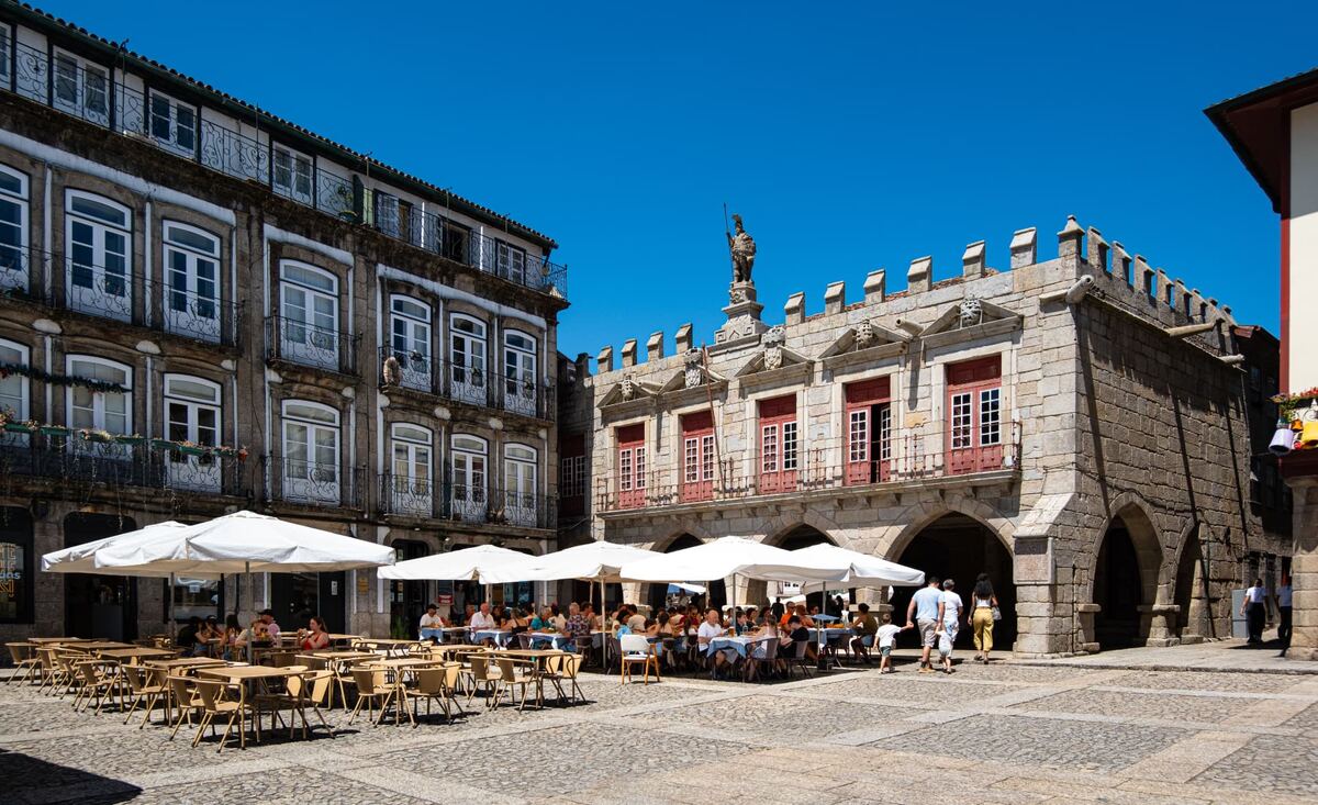 Praça histórica com esplanadas, edifícios de pedra e pessoas reunidas ao ar livre